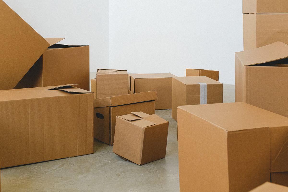 A simply furnished first apartment bedroom with a made bed, small nightstand with a lamp, and moving boxes being unpacked in the corner
